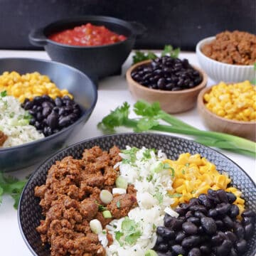 A white surface with two black bowls filled with taco meat, white rice, corn, black beans and cheese, with little bowls in the background filled with the same ingredients.