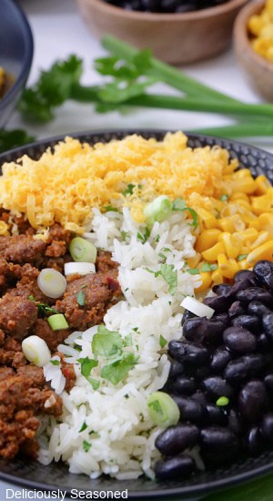 A close up of a black bowl filled with taco meat, white rice, black beans, corn and cheese.