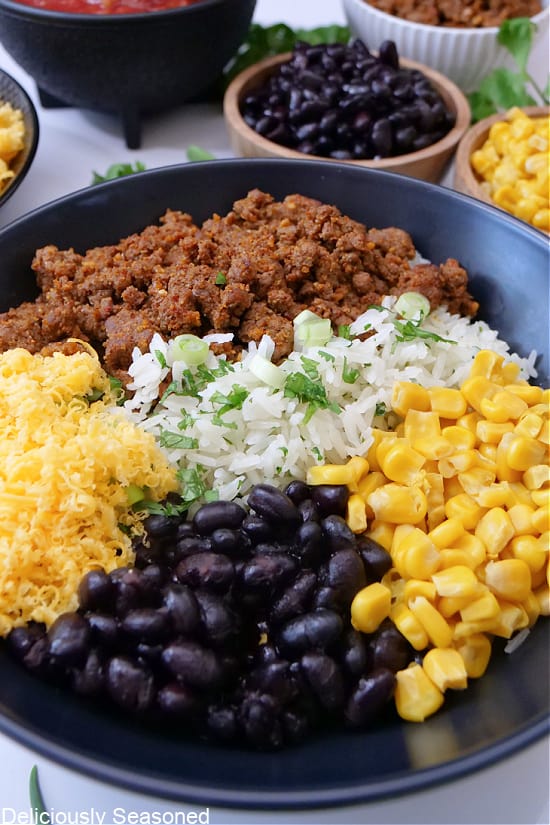 A close up photo of a black bowl with black beans, corn, rice, shredded cheese, and taco meat.