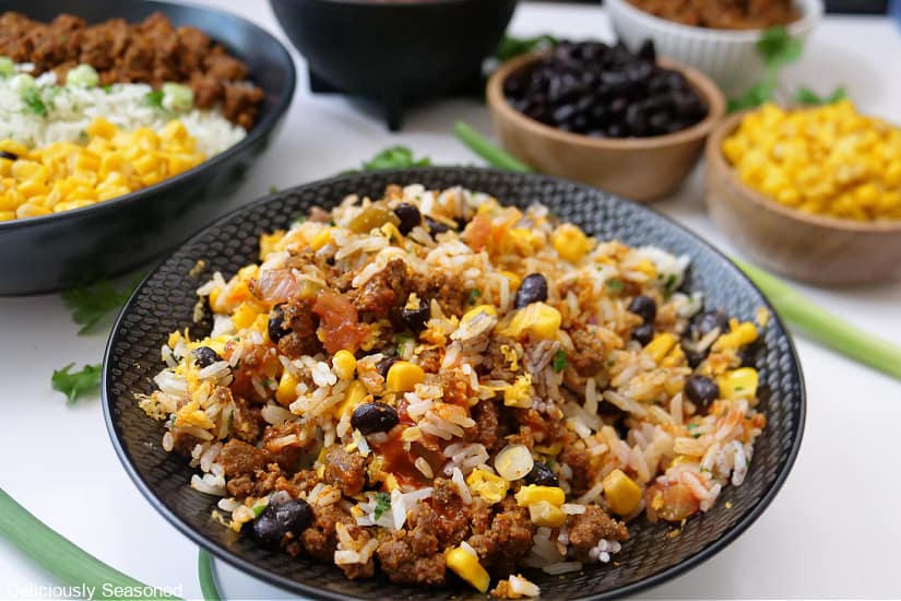 A horizontal photo of two black bowls filled with corn, rice, black beans, cheese, and taco meat.