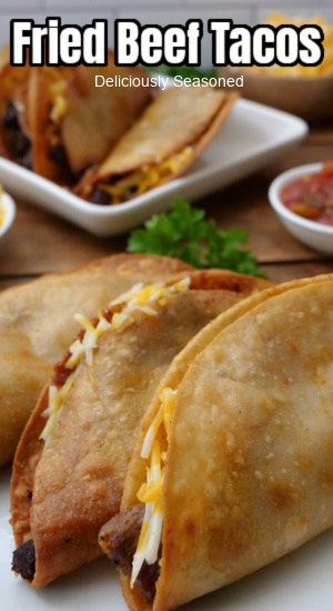 Three fried tacos on a white plate with additional tacos in the background.