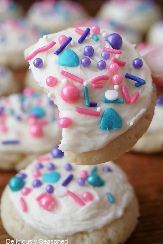 A close up photo of a sugar cookie with white buttercream frosting and candy sprinkles on it with a bite taken out of the cookie.