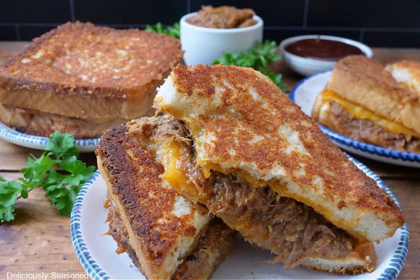 Three pulled pork grilled cheese sandwiches on plates on a table with a bowl of bbq sauce and more pulled pork in the background.