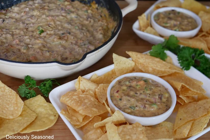 A wood surface with a white cast iron pan filled with smoked brisket queso, and two white plate and white bowls with queso and chips o\in them.
