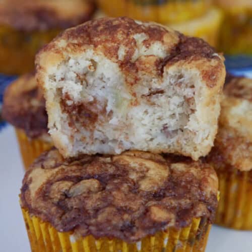 A few banana muffins on a white round plate with blue trim with one muffin on top of another, and a bite taken out of it.