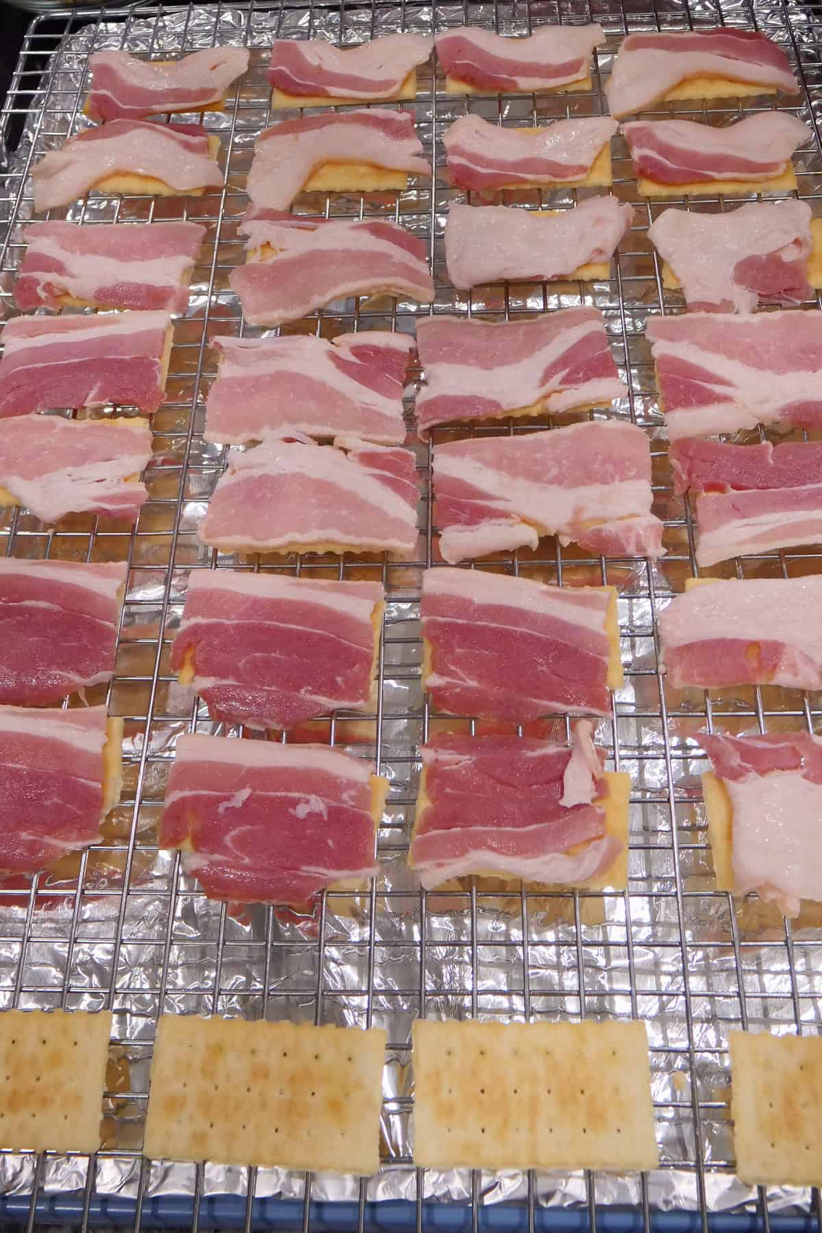 A foil lined baking sheet with a wire rack with crackers and pieces of bacon on each cracker.