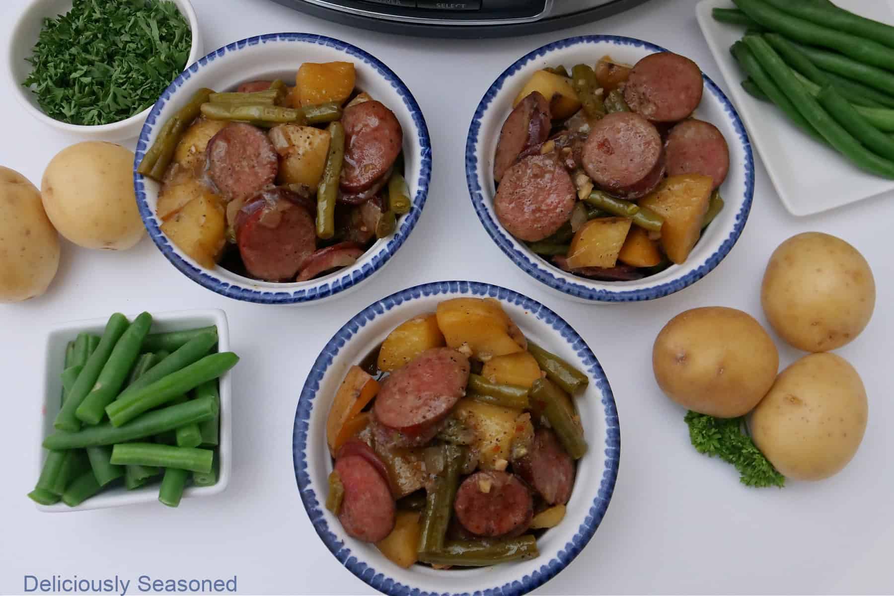 A white surface with 3 white bowls with sausage, green beans and potatoes in them.