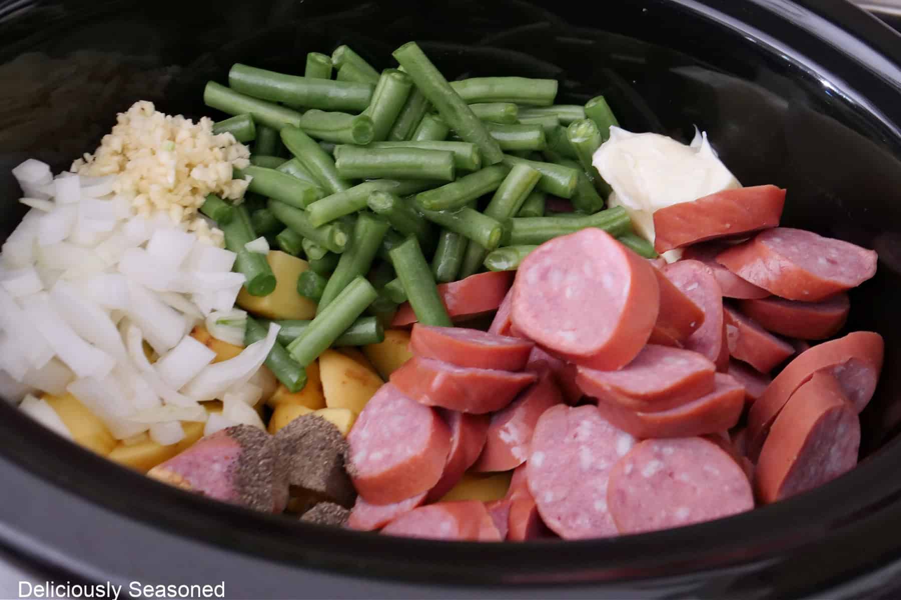 A black crock pot with all the ingredients in it to make potatoes, green beans, and sausage.