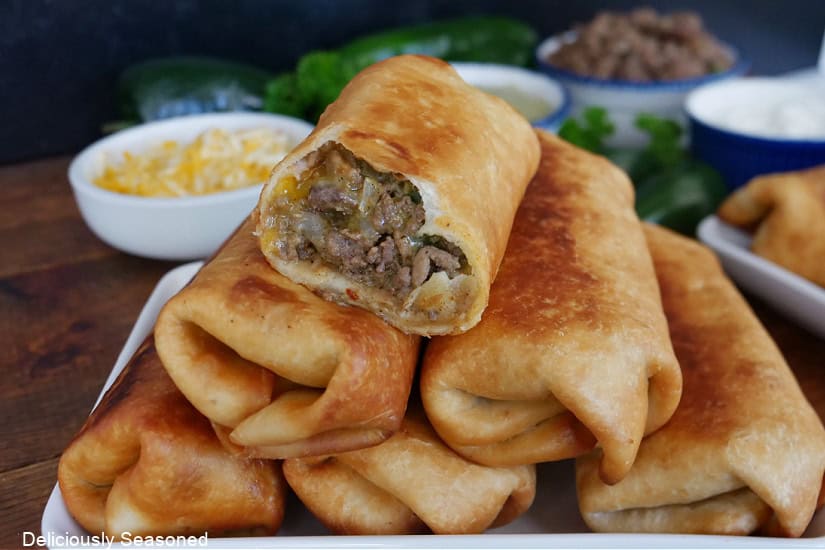 A stack of six fried beef burritos on a white plate with a few bites out of the one on top.