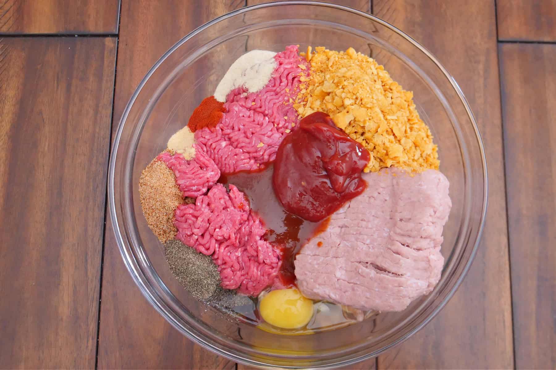A wood surface with a glass bowl filled with the ingredients to make mini brown sugar meatloaf.