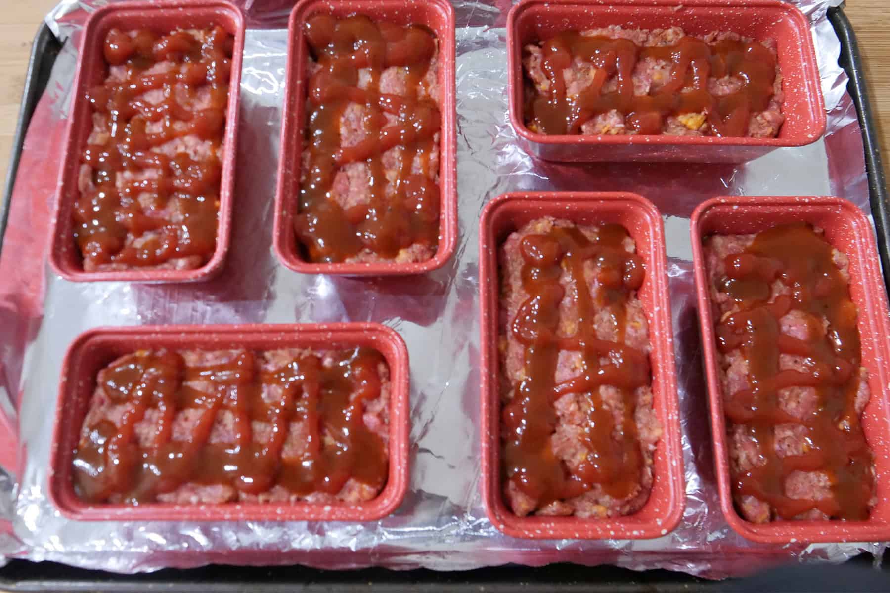 A foil lined baking sheet with six mini loaf pans filled with brown sugar meatloaf before being cooked.