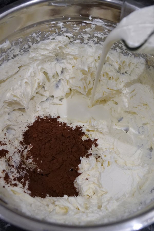A silver bowl with cream cheese, dark cocoa powder, and milk being poured into the bowl.