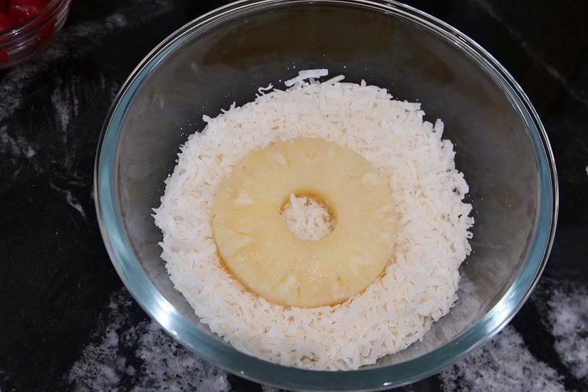 A black surface with a glass bowl with a pineapple ring being dipped into coconut.