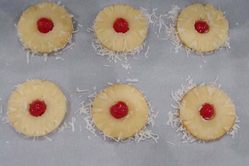 A baking sheet with six pineapple rings and cherries in the middle of each ring.