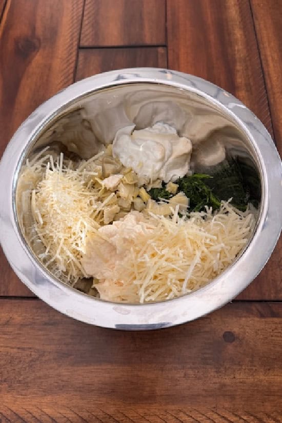 A wood surface with a silver bowl filled with ingredients to make spinach artichoke cheese bread.