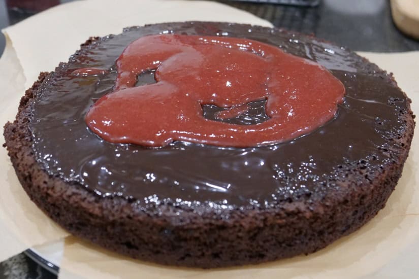 A round chocolate cake with chocolate sauce and strawberry filling spread over the top.