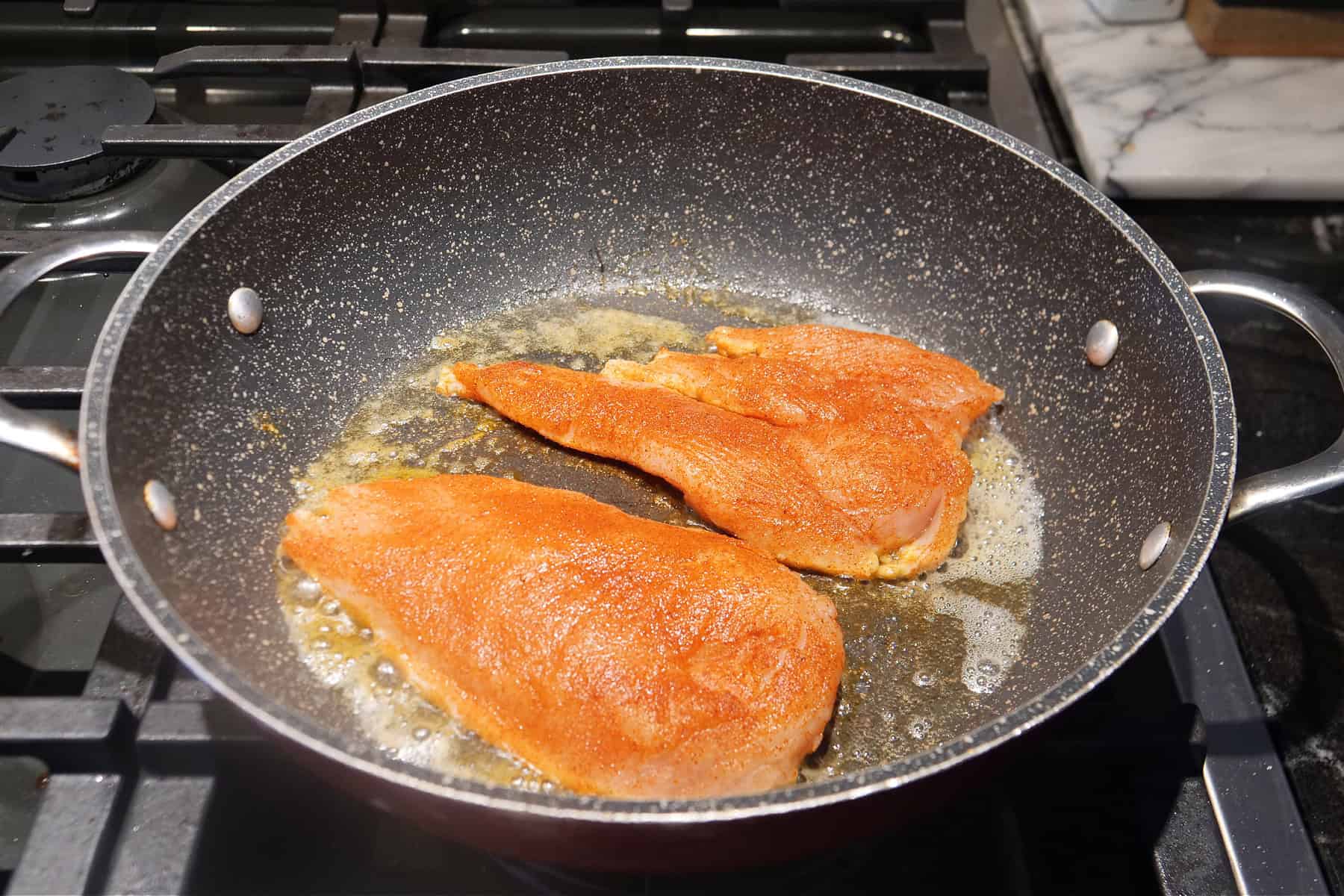 A deep skillet with two pieces of chicken breasts in it being cooked.