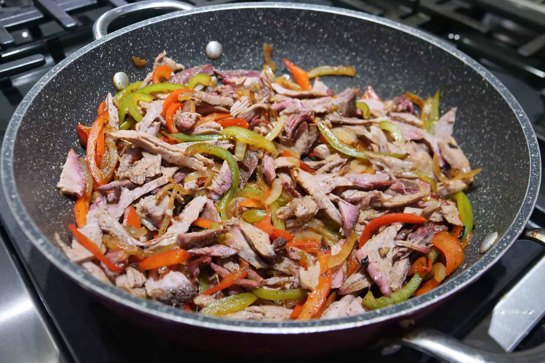 A frying pan with brisket, sliced onions, red, and green bell peppers in it.