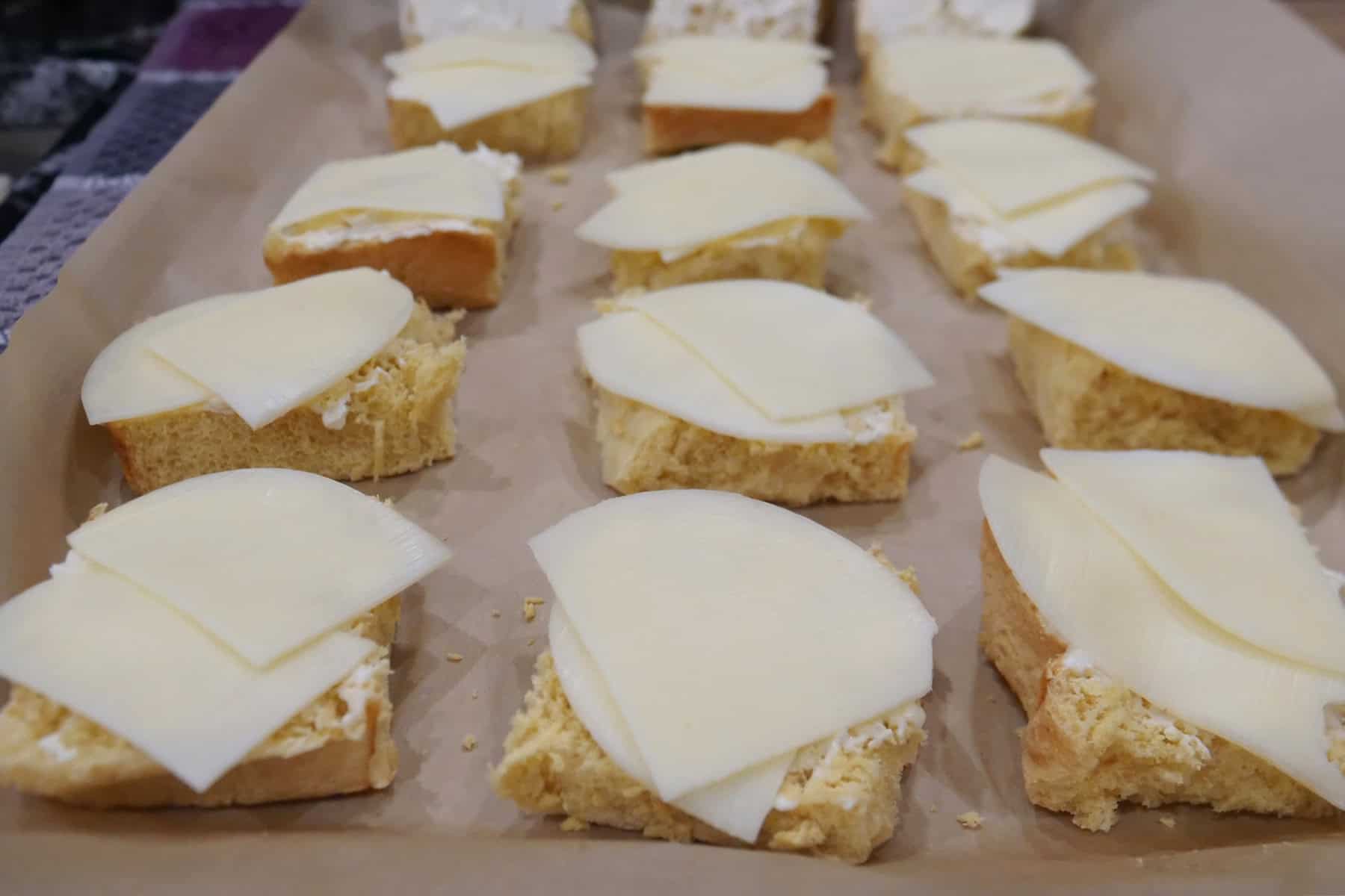 A baking sheet with 12 slider buns with cheese on them.