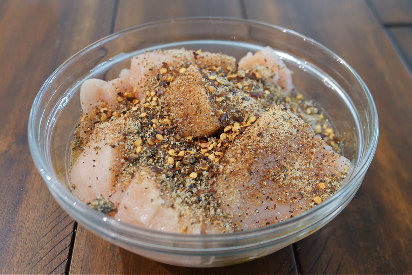 A glass bowl filled with cut chicken pieces, seasonings, and oil.