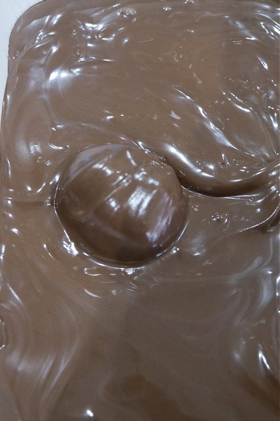A tray of melted chocolate with a single cookie butter dough ball covered with the melted chocolate.