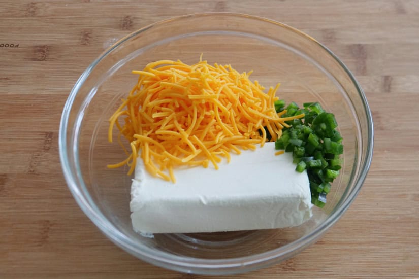 A wood surface with a glass bowl with cream cheese, shredded cheddar, and diced jalapenos in it.