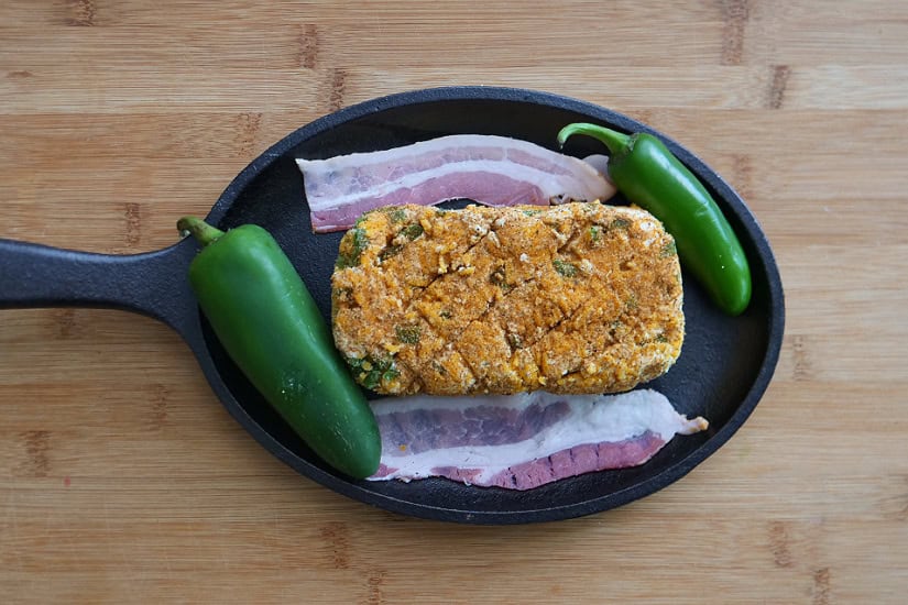 A wood surface with an oval small cast iron skillet with a brick of cream cheese mixture, two jalapenos, and two strips of bacon in it before it gets smoked.