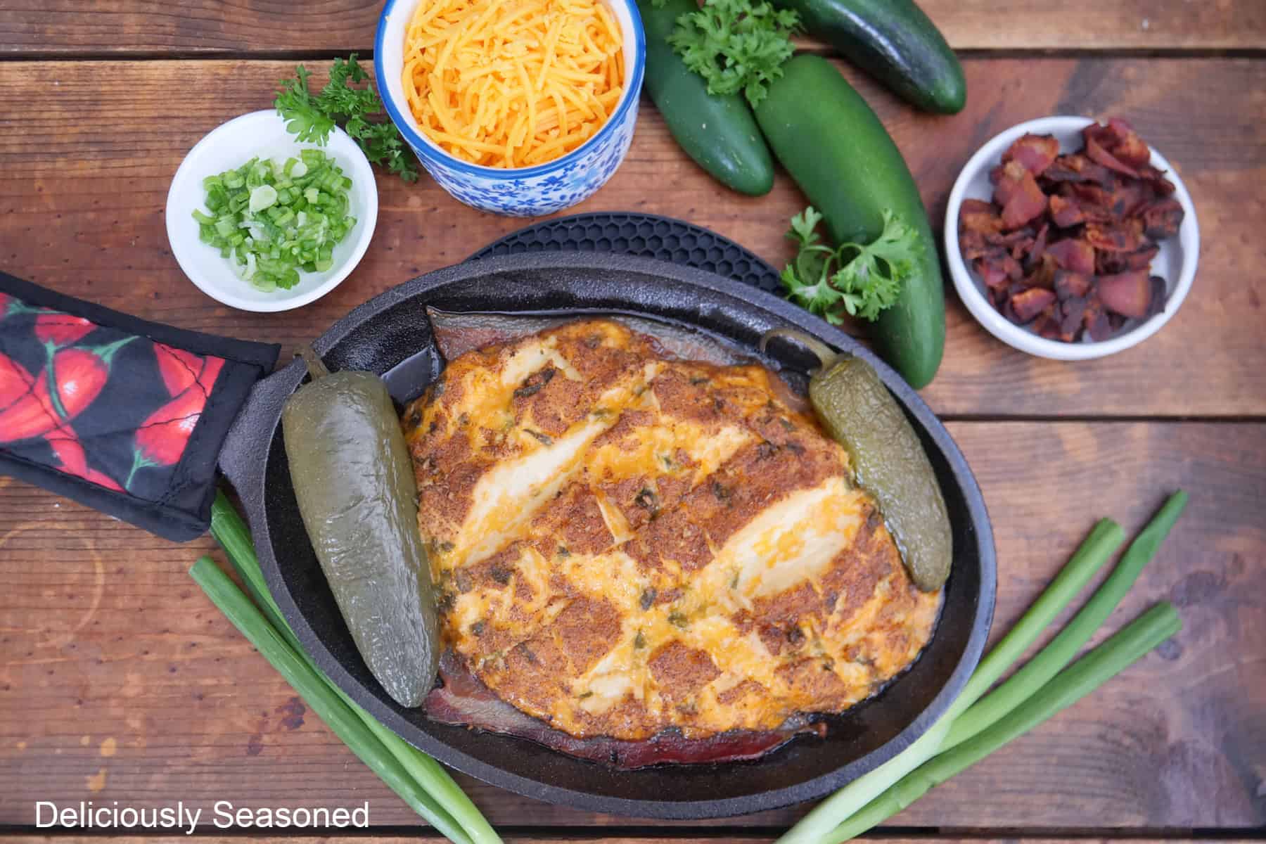 A wood surface with a small black oval cast iron skillet with smoked jalapeno popper cream cheese in it with copped bacon, green onions, and shredded creese in separate bowls.
