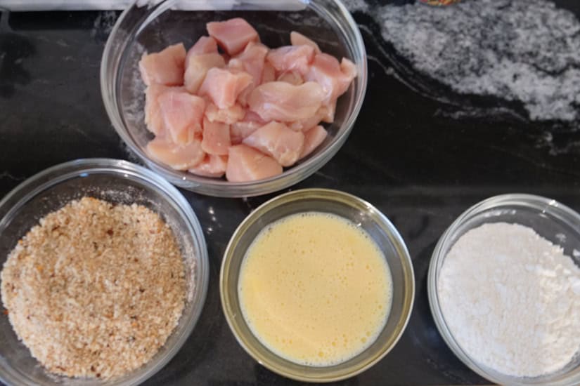 A black granite counter with 4 glass bowls: one with chicken pieces, one flour, one egg mixture, and one filled with seasoning and breadcrumbs.