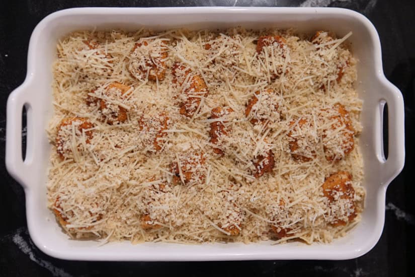 A white baking dish with pasta in a creamy sauce with fried chicken on top and covered with shredded cheese.
