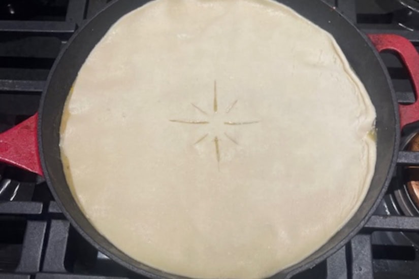 A red cast-iron pan with a pie crust with slits in the top covering up and pot pie ingredients underneath before it is baked.