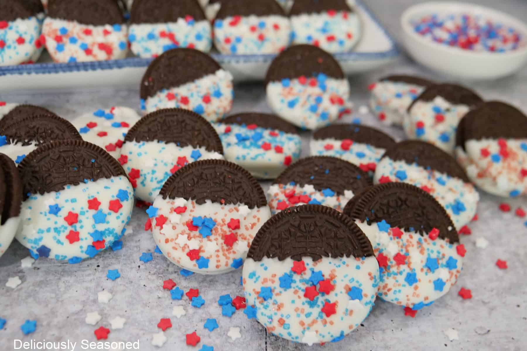 A light grey surface with a bunch of half dipped white chocolate Oreo cookies with red, white, and blue festive sprinkles on them.