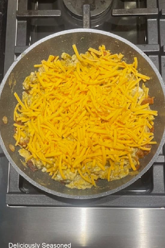 A frying pan with shredded sharp cheddar cheese sprinkled over scrambled eggs and crumbled bacon.