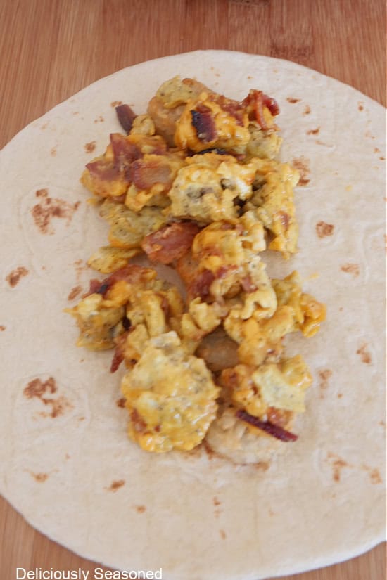 A wood surface with a flour tortilla on it with eggs, bacon, cheese, salsa on it.
