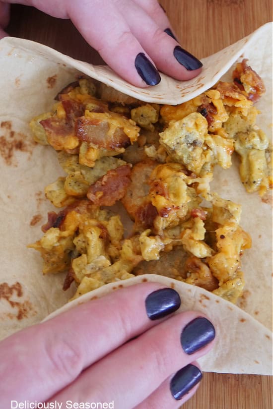 Two hand rolling up a flour tortilla filled with eggs, bacon, cheese, and salsa, into a burrito.