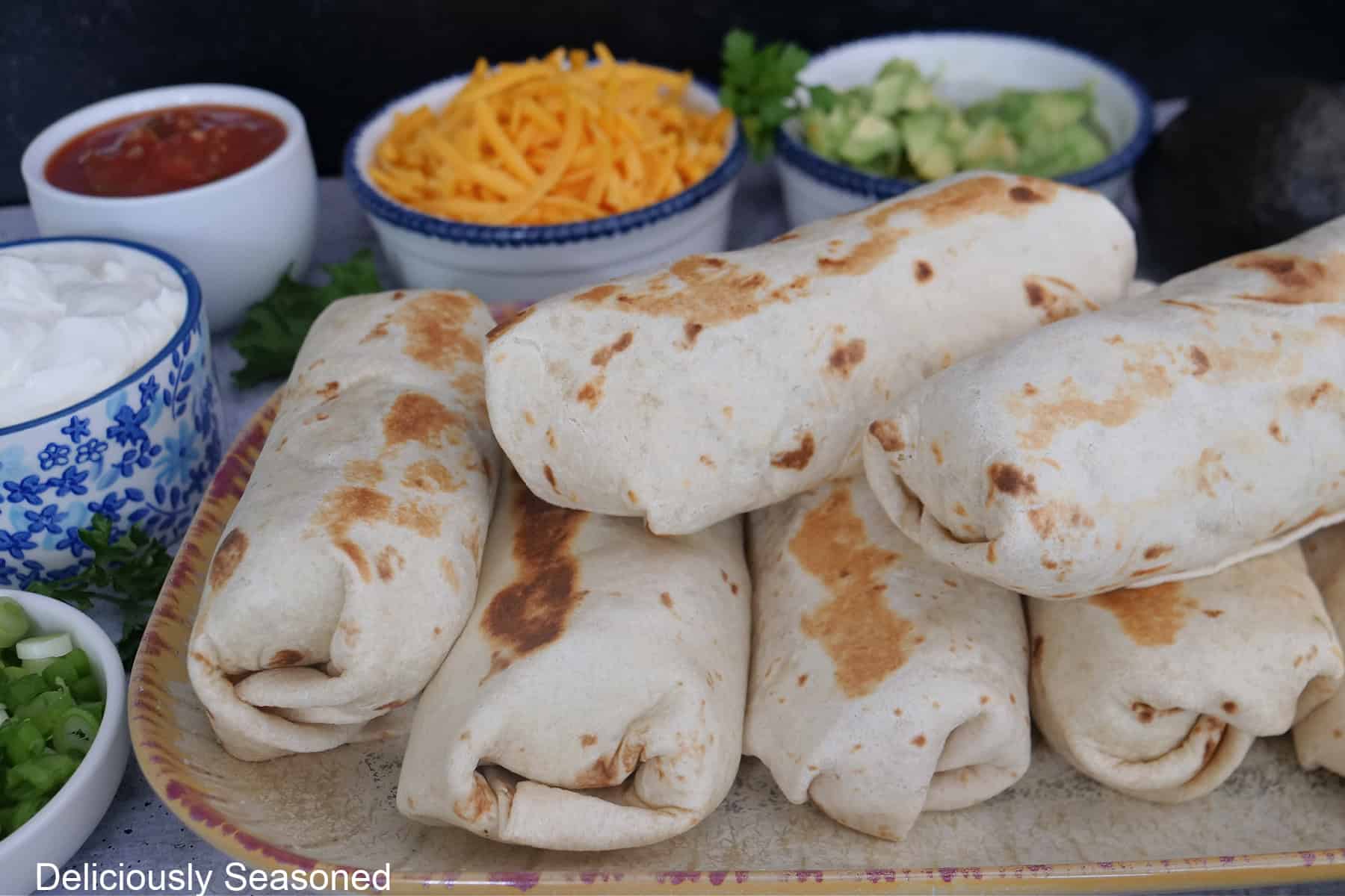 A beige oblong plate with six breakfast burritos on it with small bowls filled with different toppings in the background.