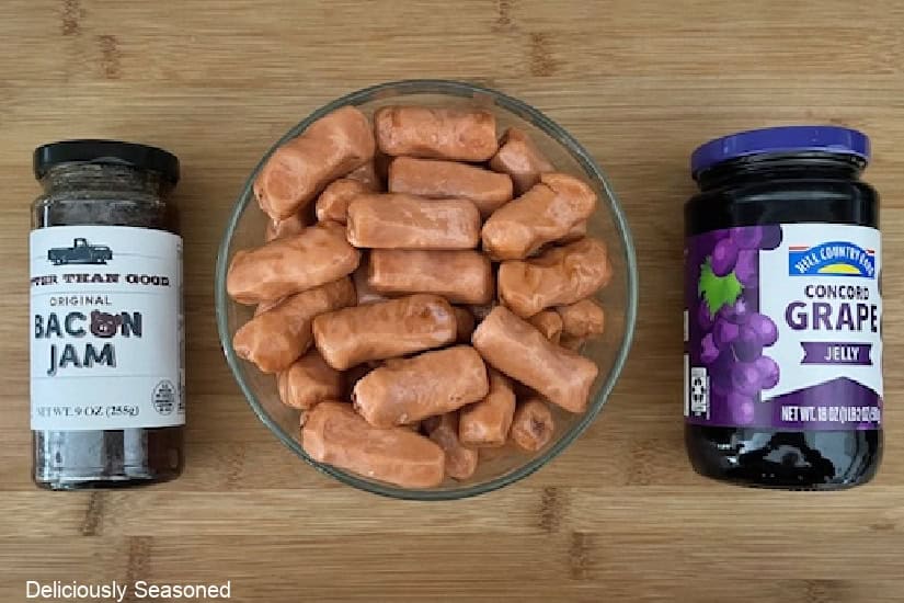 A wood surface with a glass bowl filled with little smokies, a jar of bacon jam and a jar of grape jelly.