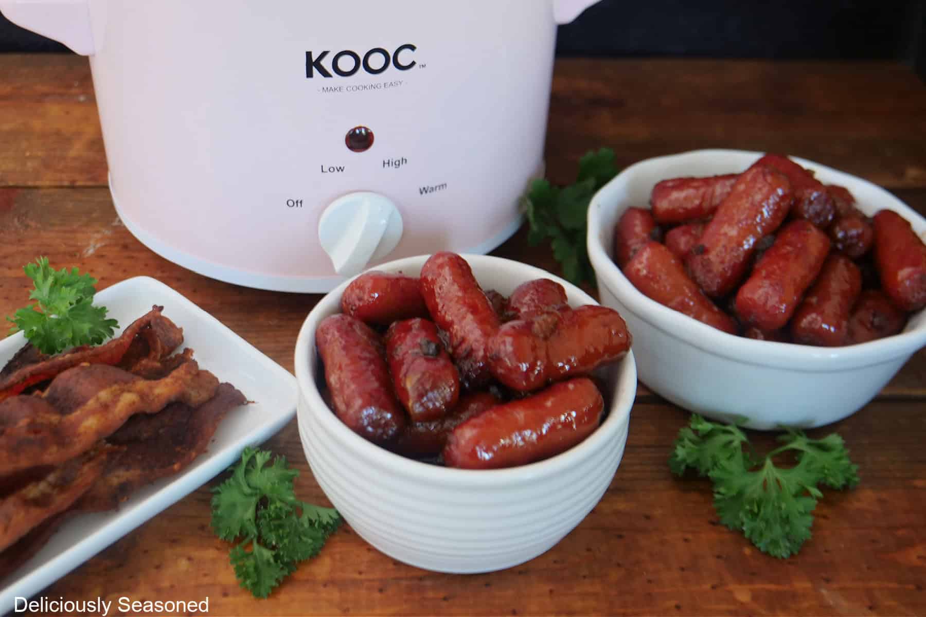 A wood surface with two white bowls filled with little smokies, and a white plate with cooked bacon on it, and a small crockpot in the background.