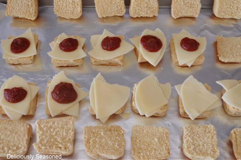 A foil lined baking sheet with slider buns on it topped with pizza sauce and cheese.