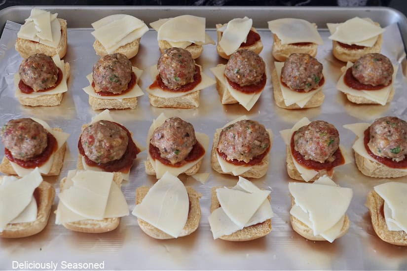 A foil lined baking sheet with slider buns on it with sliced cheese, pizza sauce, and turkey meatballs.