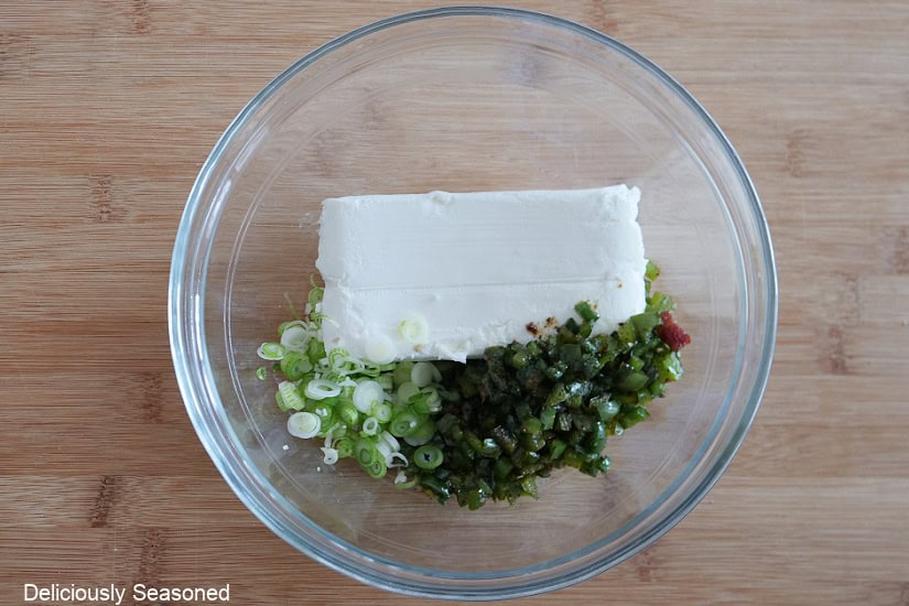 A glass bowl with cream cheese, diced jalapenos, green onions, and seasoning in it.