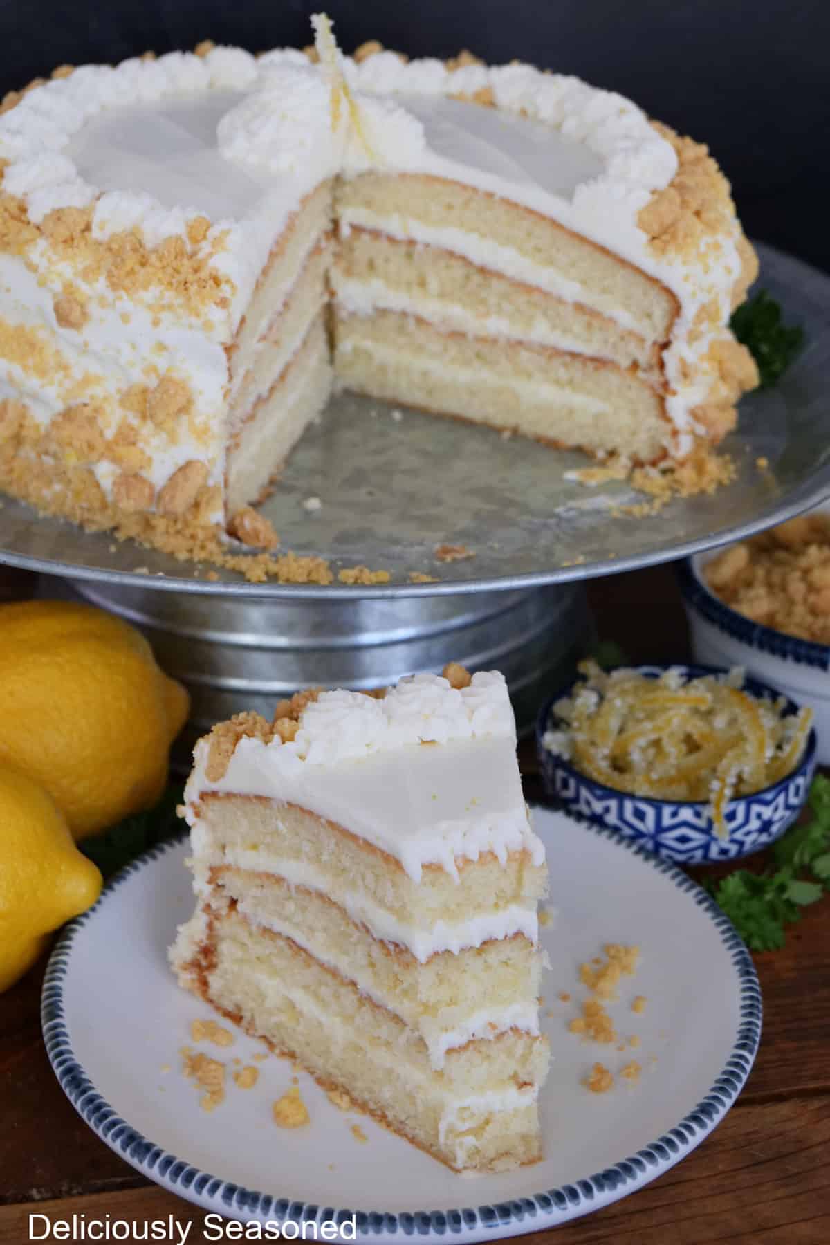 A slice of lemon crunch layer cake on a white plate with blue trim and a silver cake plate with the whole lemon layered cake on it with a slice taken out of it.
