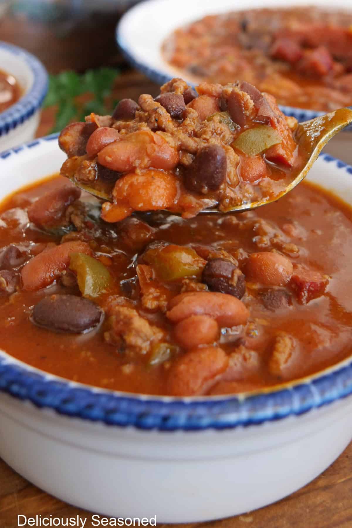 A white bowl with blue trim with a serving of homemade chili in it and a spoonful of chili held above the bowl.