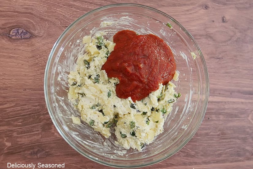 A glass mixing bowl with a cream cheese mixture combined together and pizza sauce added to the bowl.