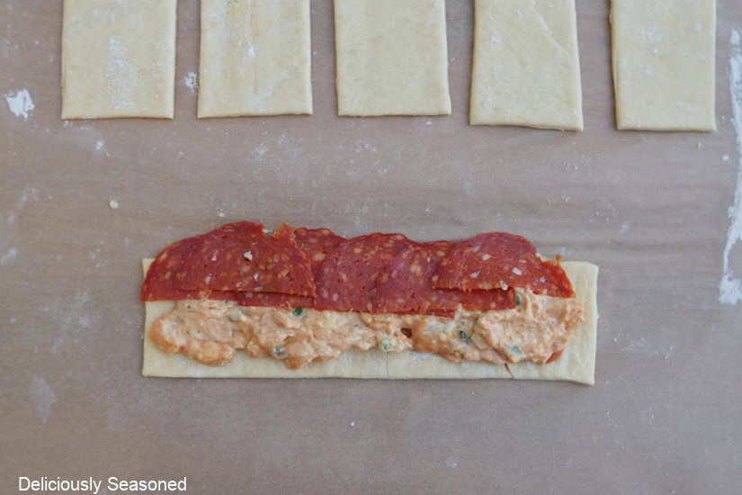 A strip of crescent dough with cream cheese mixture and half slices of pepperoni placed on it before being rolled into a pinwheel.