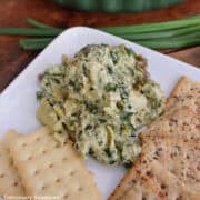 A square white plate with spinach artichoke pesto dip on it with crackers on both sides of the dip.