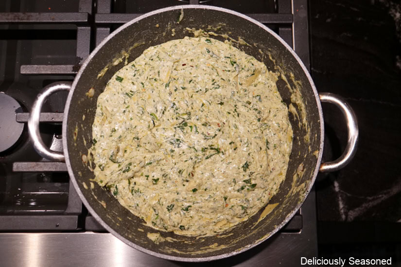 A frying pan with all the ingredients mixed together to make pesto spinach artichoke dip in it.