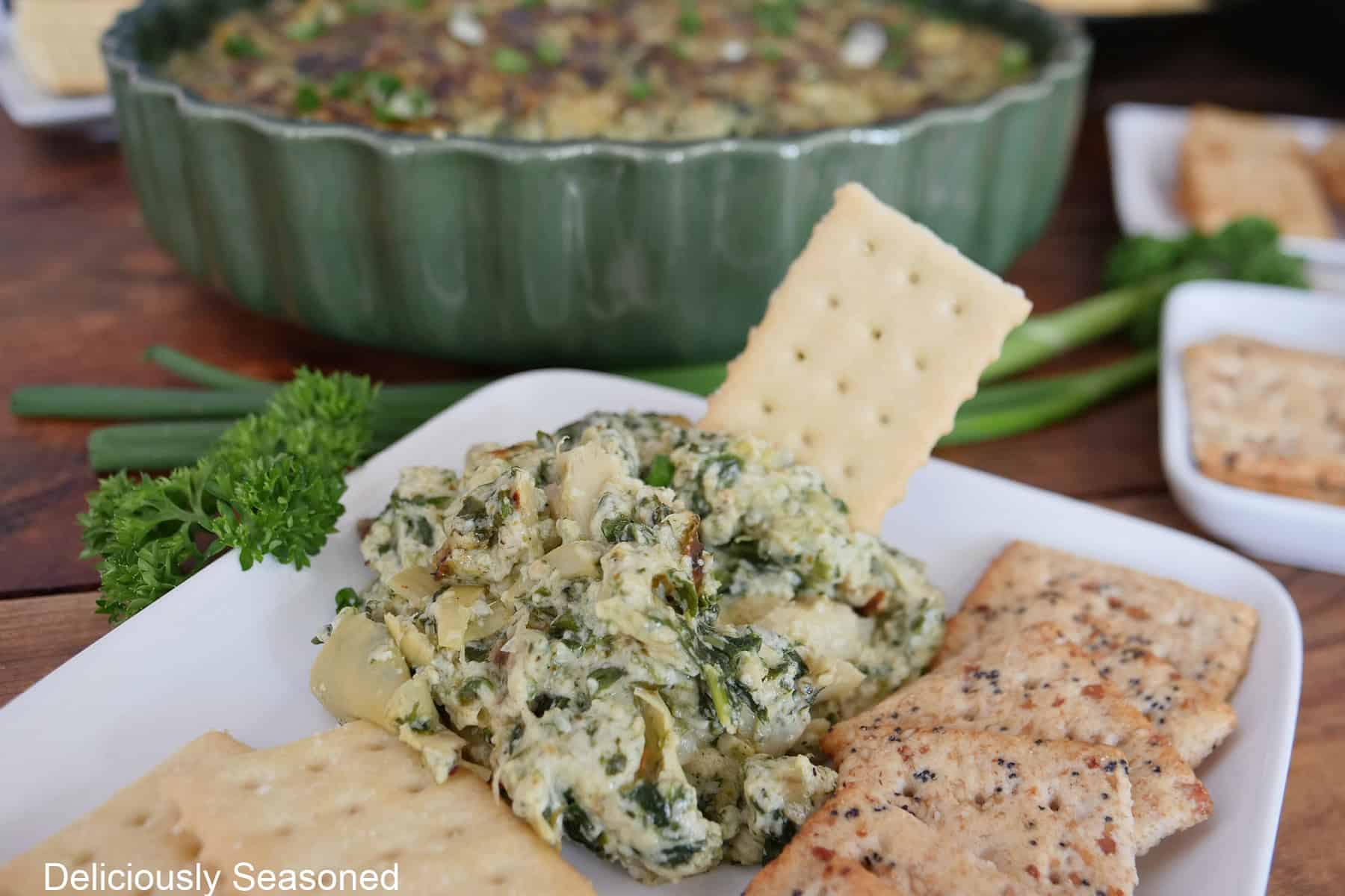 A white plate with crackers and some pesto spinach and artichoke dip on it.