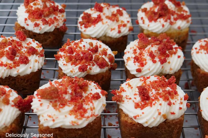 A wire rack with mini cheesecake bites on it that have been topped with whipped cream and a raspberry crunch topping.