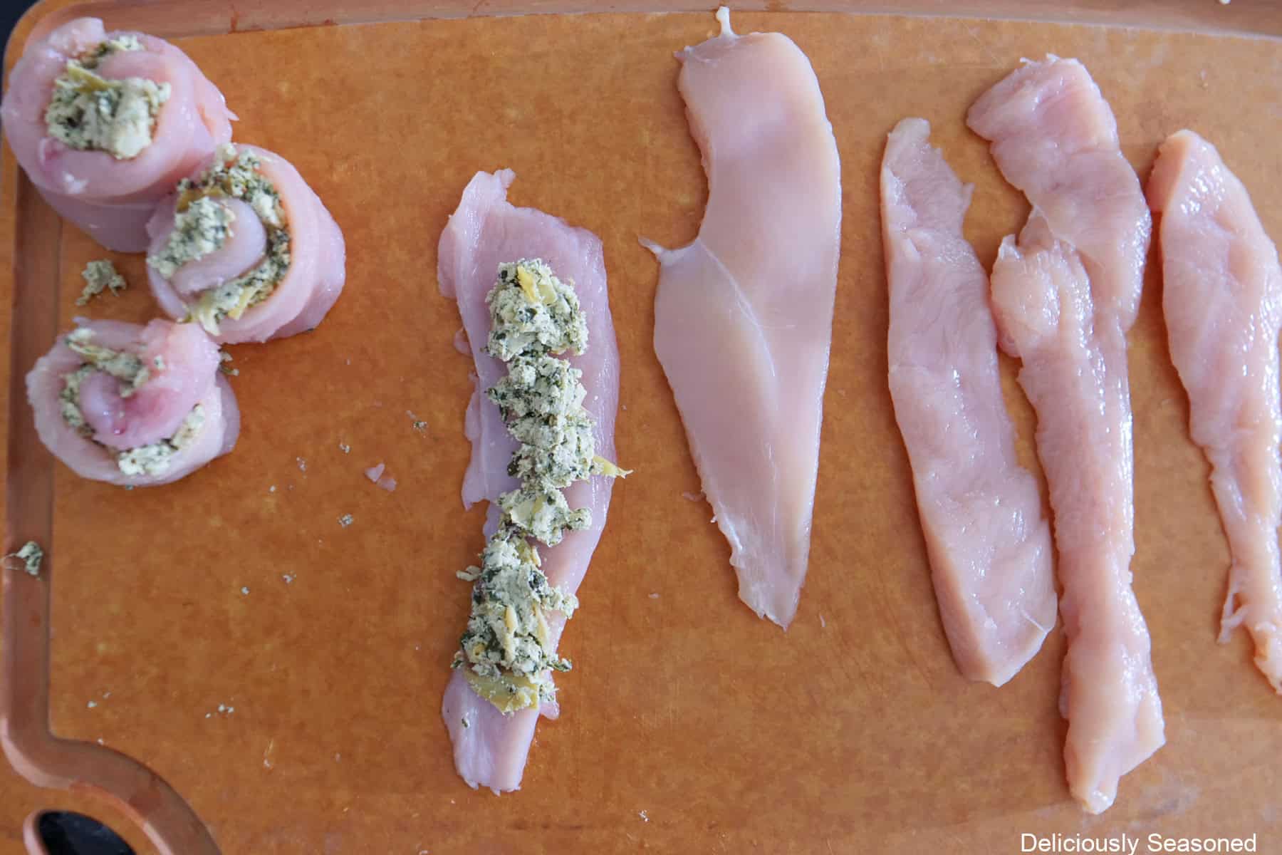 A wood cutting board with strips of raw chicken breasts cut into 2 inch strips and some are stuffed and rolled up, one is topped with spinach artichoke dip, with four more strips placed on the cutting board.